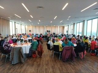 Ein großer Raum mit Holzböden und großen Fenstern, in dem viele Menschen um Tische herum sitzen, wahrscheinlich in einer Konferenz oder Besprechungssituation. Einige tragen bunte Schals.