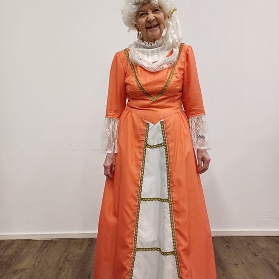 Eine ältere Frau in einem orangefarbenen und weißen Kleid mit weißem Spitzenkragen und goldener Einfassung. Sie hat weißes Haar und lächelt.