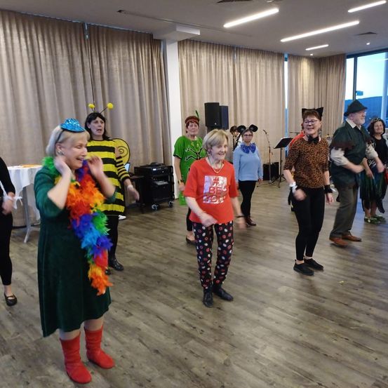 Eine Gruppe von Erwachsenen in Kostümen tanzt in einem Raum mit Holzböden, einer trägt ein Bienenkostüm. Im Hintergrund stehen Musikinstrumente und ein Tisch.