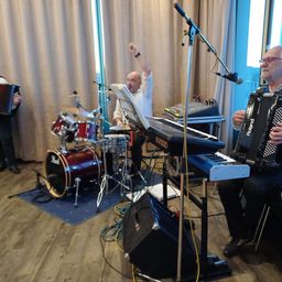 Eine Gruppe von Musikern tritt auf der Bühne auf; ein Schlagzeuger, ein Keyboarder und zwei Akkordeonisten. Der Schlagzeuger spielt an einem Schlagzeug mit Becken, während der Keyboarder an einem Keyboard mit einem Mikrofonständer spielt.