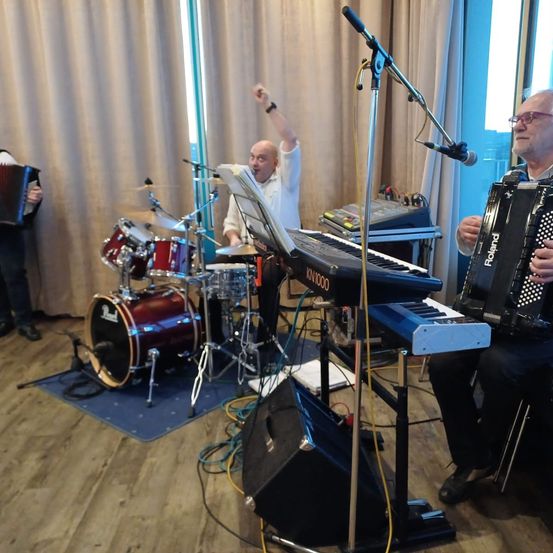 Eine Gruppe von Musikern tritt auf der Bühne auf; ein Schlagzeuger, ein Keyboarder und zwei Akkordeonisten. Der Schlagzeuger spielt an einem Schlagzeug mit Becken, während der Keyboarder an einem Keyboard mit einem Mikrofonständer spielt.