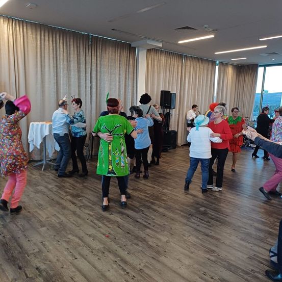 Eine Gruppe von Menschen in festlicher Kleidung tanzt in einem Raum mit Holzböden, großen Vorhängen und einem Lautsprecher.