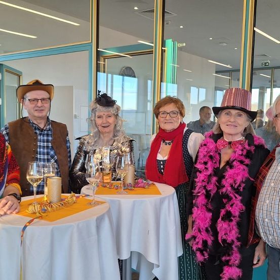 Eine Gruppe von Menschen in Kostümen lächelt in die Kamera und steht um Tische mit Weingläsern herum. Hinter ihnen führen Glastüren zu einem helleren Raum.