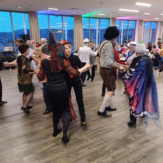 Eine Gruppe älterer Menschen tanzt in einem Saal mit Holzböden, einige in Kostümen. Andere sitzen um sie herum. Der Saal hat große Fenster.