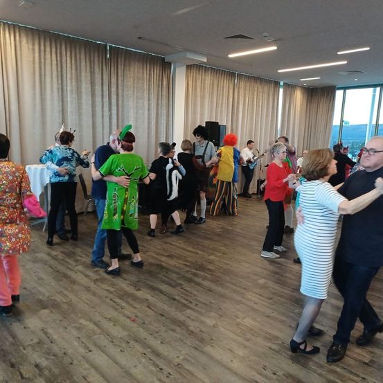 Eine Gruppe von Menschen tanzt in einem Raum mit Holzboden. Einige tragen Kostüme und Brillen. Ein Tisch mit weißer Tischdecke steht links. Ein Mann spielt Gitarre. Vorhänge und Lampen sind an der Wand.