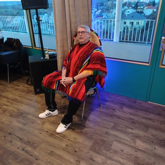 Eine ältere Frau in einem bunten Poncho sitzt auf einem Stuhl in einem Raum mit Holzböden. Hinter ihr befindet sich ein Fenster mit Blick auf Häuser und einen Balkon.