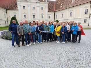 Eine Gruppe von Menschen in Jacken und Turnschuhen posiert für ein Foto vor einem historischen Gebäude mit roten Dächern und gepflastertem Boden.