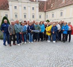 Eine Gruppe von Menschen in Jacken und Turnschuhen posiert für ein Foto vor einem historischen Gebäude mit roten Dächern und gepflastertem Boden.
