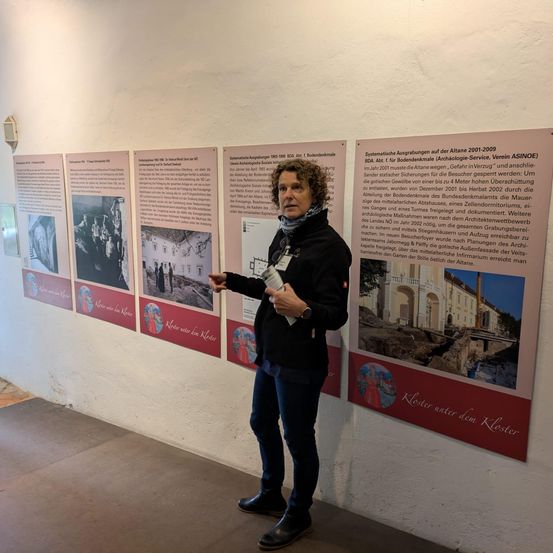 Eine Frau steht vor einer Wand mit Informationspostern, die die Geschichte und Restaurierung eines alten Gebäudes erklären. Sie hält ein Stück Papier in der Hand und zeigt auf die Poster.