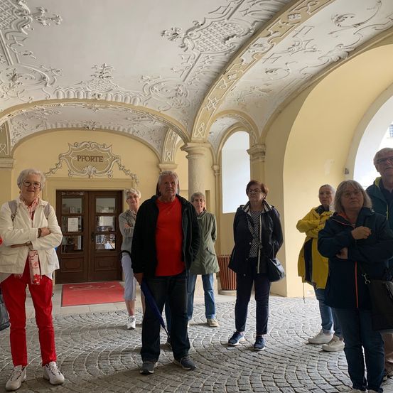 Eine Gruppe von Menschen in einem historischen Flur mit dekorativen Decken. Sie stehen herum und plaudern, gekleidet in warme Kleidung. Der Eingang zu einem Raum ist im Hintergrund zu sehen.