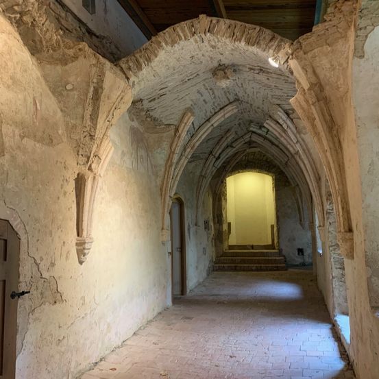 Ein schmaler, alter Bogengang mit Ziegelboden und Steinmauern. Am Ende scheint Licht durch.