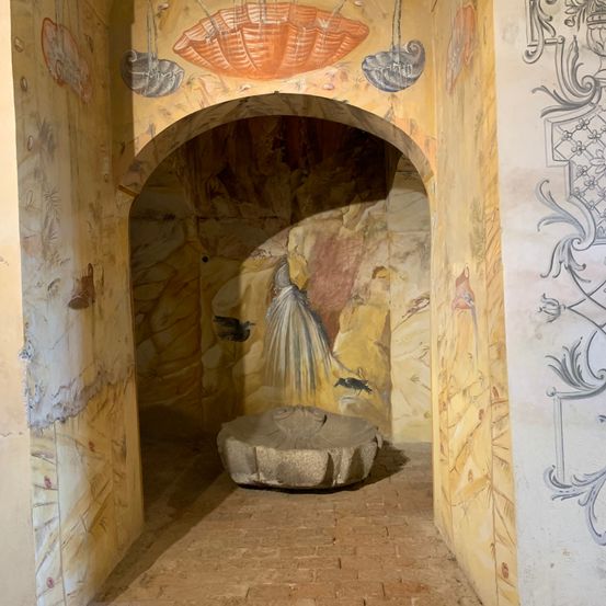 Ein Bogen führt in einen Raum mit bemalten Wänden, die eine Frau mit einem fließenden Kleid und Vögeln darstellen. Ein Steinbecken steht auf dem Boden.