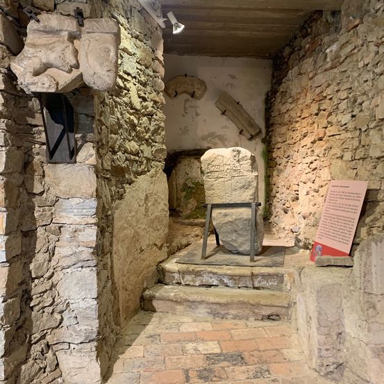 Ein Steinraum mit einem großen Felsen in der Mitte, umgeben von Steinmauern und Stufen, die zu ihm hinaufführen. Ein Metallrahmen umgibt den Felsen, und eine Lampe ist an der Decke montiert.