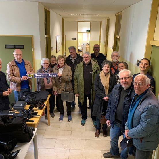 Eine Gruppe von Menschen steht in einem Flur, einer hält ein blaues Banner mit der Aufschrift 'Senioren Horn'. Verschiedene Taschen und Gegenstände liegen auf dem Tisch. Sie alle lächeln und scheinen für ein Foto zu posieren.