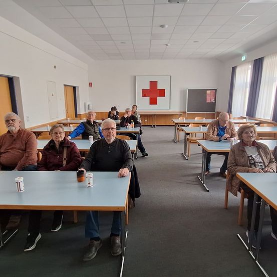 Eine Gruppe von Menschen sitzt an Tischen in einem Raum mit einem roten Kreuzschild an der Wand. Es gibt Fenster mit Vorhängen, und einige Personen tragen Brillen und Turnschuhe.