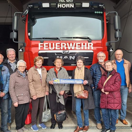 Eine Gruppe von Menschen, darunter ältere Frauen, steht vor einem roten Feuerwehrauto. Sie lächeln und posieren für ein Foto, wobei eine Frau ein Schild mit der Aufschrift 'Senioren Horn' hält.