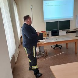 Ein Mann in Feuerwehruniform steht in einem Klassenzimmer und präsentiert vor einer Projektionsleinwand. Die Leinwand zeigt ein Datum, 'Gegründet 1873'. Verschiedene Gegenstände wie Laptop, Kabel und ein Telefon sind auf dem Tisch.