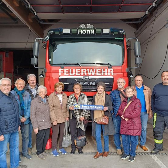 Eine Gruppe älterer Menschen steht vor einem roten Feuerwehrauto, lächelt und posiert für ein Foto. Einige halten ein Schild mit der Aufschrift 'Senioren Horn'. Der Ort ist eine Feuerwache.