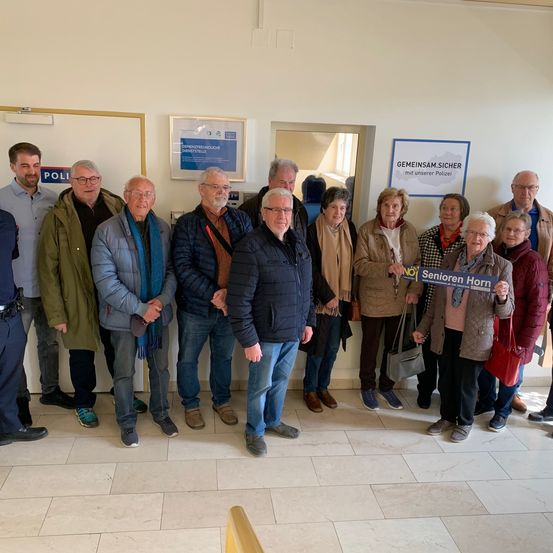 Eine Gruppe älterer Erwachsener steht zusammen in einem Raum mit der Polizei und hält ein Schild mit der Aufschrift 'Senioren Horn.' Hinter ihnen befinden sich weiße Wände mit gerahmten Plakaten.
