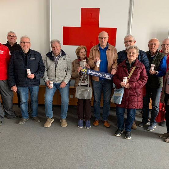 Eine Gruppe älterer Menschen, alle mit Tassen in der Hand, steht vor einem roten Kreuz an einer weißen Wand. Sie lächeln und posieren für ein Foto.