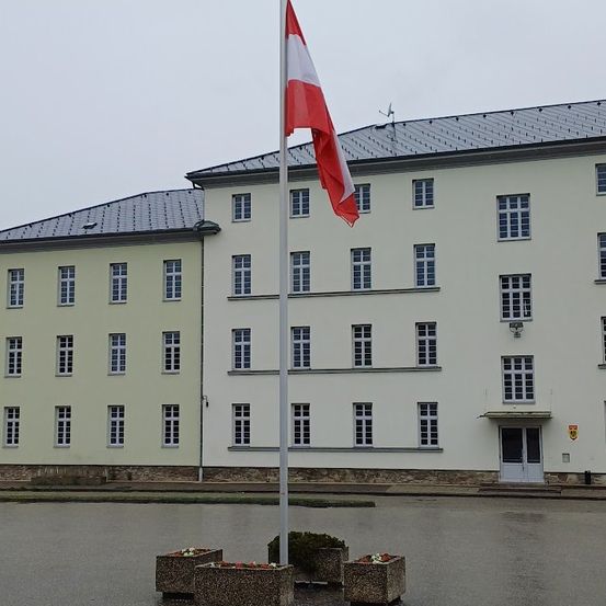 Eine rot-weiße Flagge steht vor einem großen Gebäude mit vielen Fenstern und einem flachen Dach. Das Gebäude hat einen zentralen Eingang. Vor dem Fahnenmast stehen drei Blumenkästen.