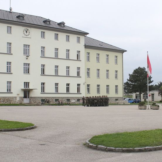 Eine Gruppe von Menschen in Militäruniformen steht in einer Reihe vor einem Gebäude. Das Gebäude hat mehrere Fenster und eine Uhr an der Wand. Vor dem Gebäude sind Fahrräder geparkt.