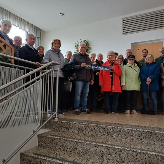Eine Gruppe von Menschen steht auf einer Treppe und hält ein blaues Schild mit 'Senioren Horn' darauf. Sie sind warm gekleidet und einige tragen Brillen.