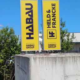 Zwei gelbe Banner, eines mit der Aufschrift 'HABAU' und das andere mit 'HELD & FRANCK', stehen auf einem Betonblock. Dahinter befinden sich einige Bäume und ein Gebäude. Der Himmel ist klar und blau.