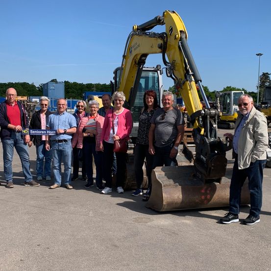 Eine Gruppe von Menschen posiert für ein Foto vor einem gelben Bagger, mit einem Banner, das 'Senioren Horn' steht. Sie sind auf einer Betonfläche mit Baumaschinen und Bäumen im Hintergrund.