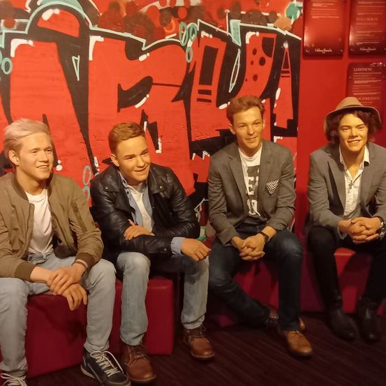 Vier junge Männer sitzen auf roten Bänken, möglicherweise für ein Fotoshooting. Sie tragen Freizeitkleidung. Hinter ihnen befindet sich eine Wand mit Graffiti. Der Mann auf der rechten Seite trägt einen Hut.