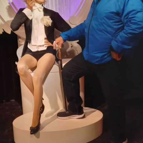 Ein Mann steht neben einer Puppe, die einen Smoking mit Fliege und Blume trägt und einen Stock hält. Er trägt eine blaue Jacke und Turnschuhe.