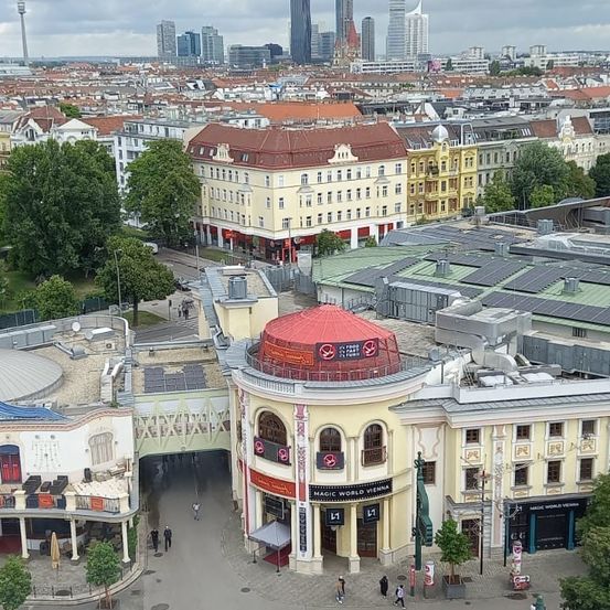 Ein Blick aus der Luft auf eine Stadt mit zahlreichen Gebäuden, einem prominenten Magic World Vienna und Menschen, die auf der Straße gehen. Die Stadt hat viele Bäume und einige hohe Gebäude in der Ferne.