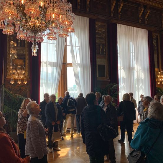 Eine Gruppe von Menschen steht in einem großen Raum mit weißen Vorhängen und einem Kronleuchter und schaut nach oben zur Decke. Der Raum ist mit kunstvollen Holzdetails und großen Fenstern geschmückt.