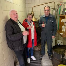 Drei Menschen stehen in einem Raum mit einem Kupferdestillierapparat. Sie halten Gläser und lächeln für ein Foto. Der Mann trägt eine Brille und eine Jacke. Die Frau trägt eine rote Jacke und eine Brille. Ein anderer Mann trägt eine grüne Arbeitskombi.