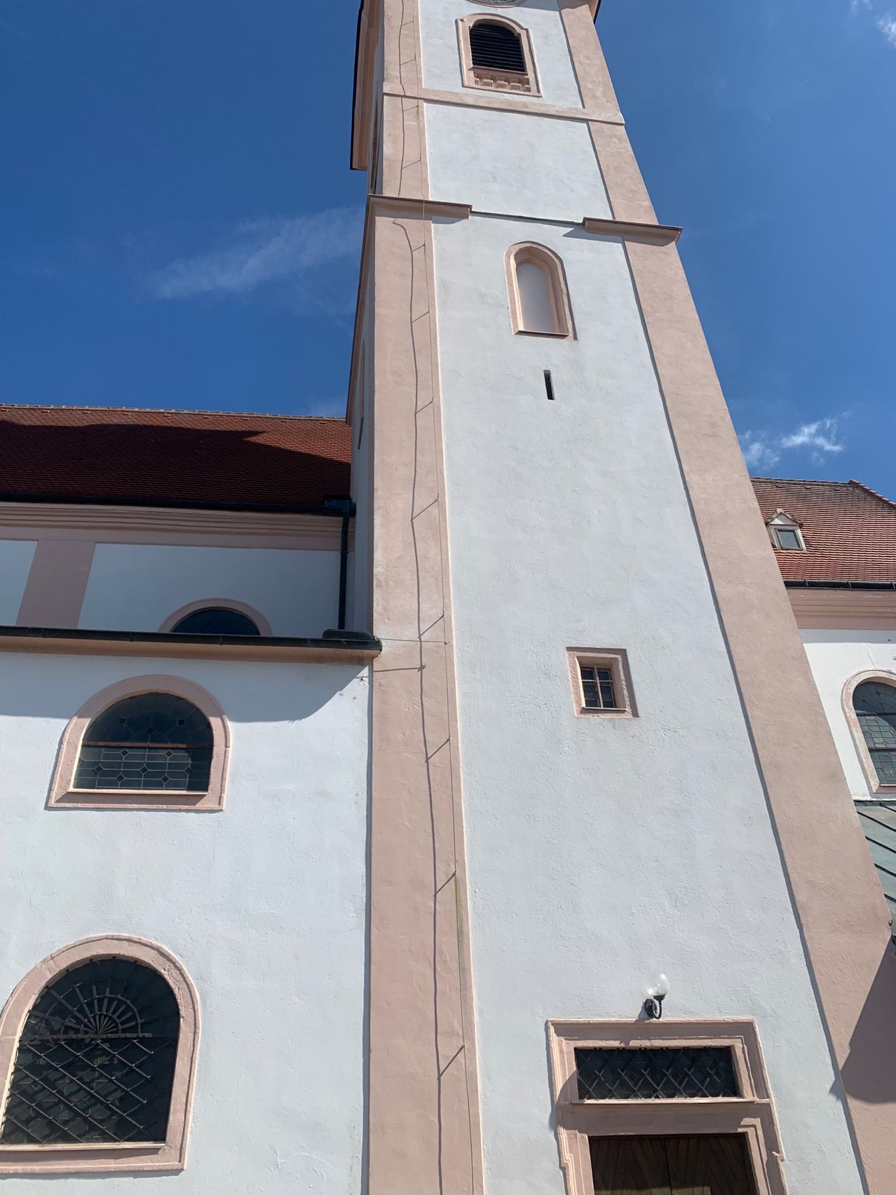 Ein hoher, weißer Kirchturm mit einem braunen Dach, gewölbten Fenstern und einer kleinen Lampe an der Seite.