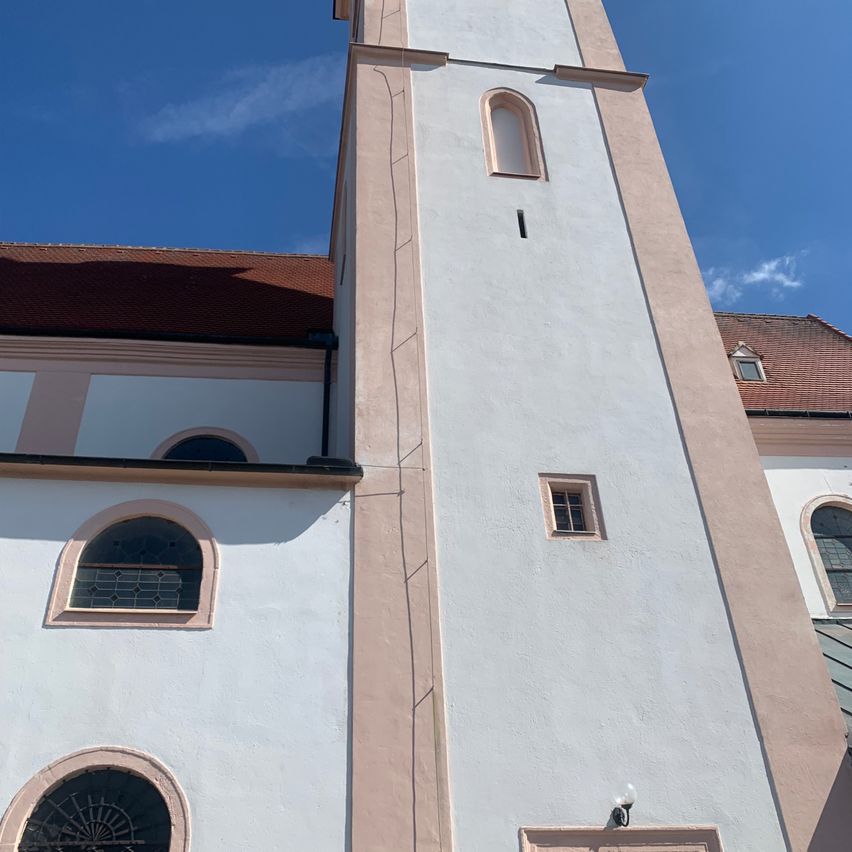 Ein hoher, weißer Kirchturm mit einem braunen Dach, gewölbten Fenstern und einer kleinen Lampe an der Seite.