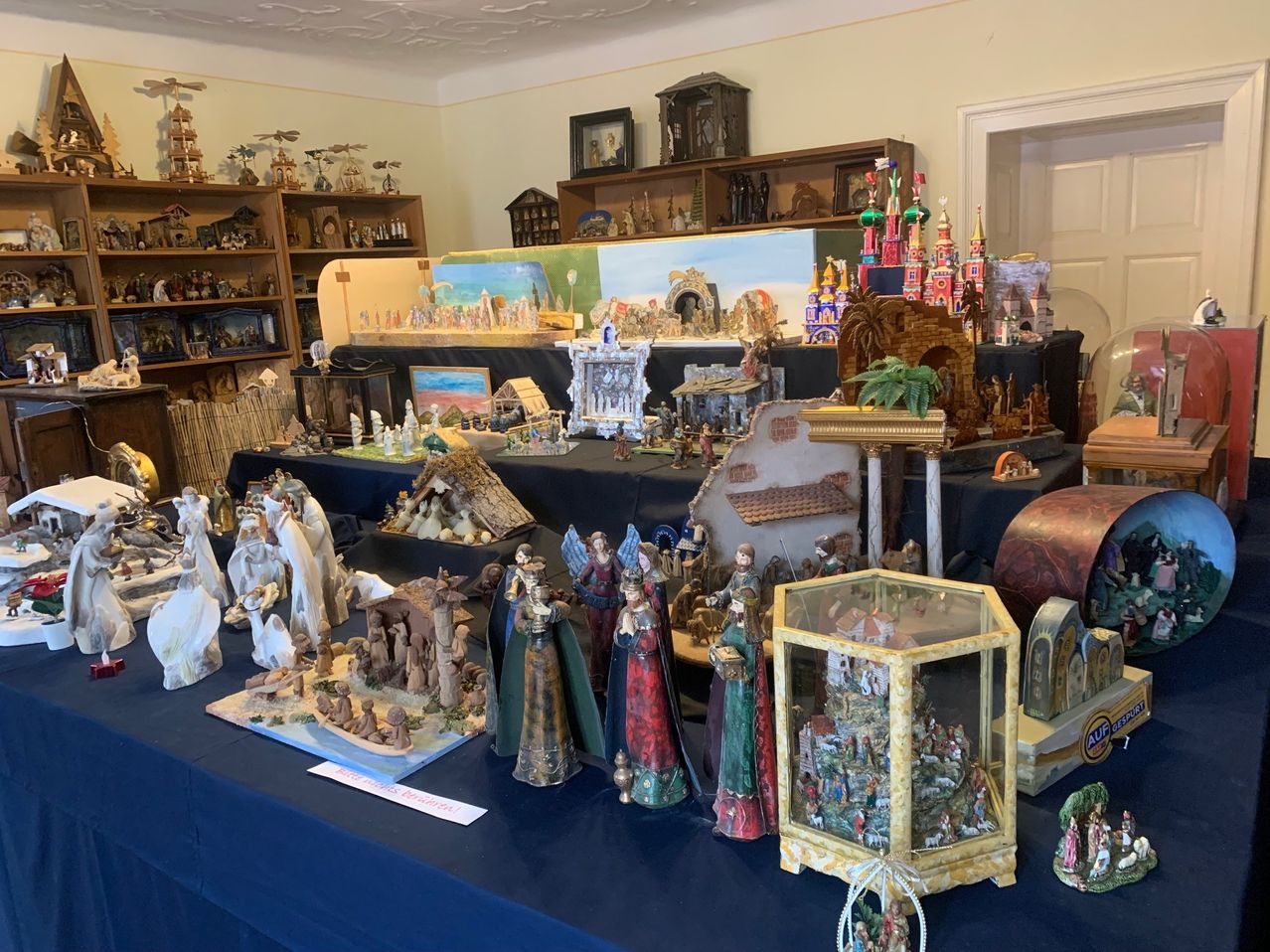 Ein Raum voller verschiedener Figuren und Skulpturen, einschließlich einer Krippe, auf einem Tisch und Regalen. Es gibt auch Bilderrahmen und Schachteln mit Weihnachtsdekoration.