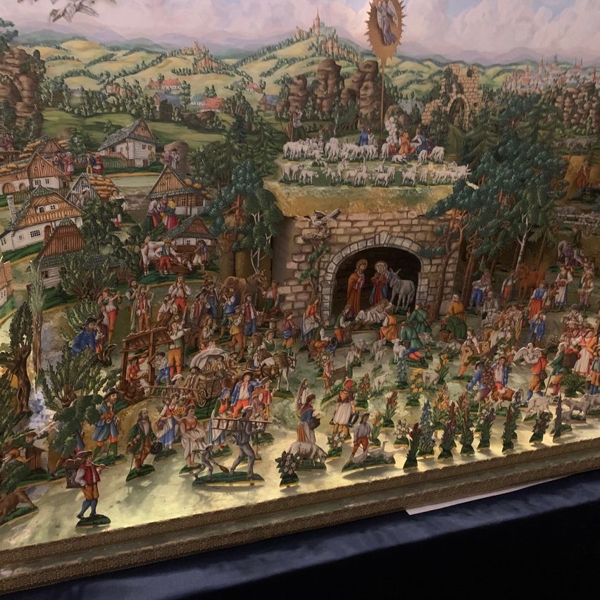 Ein detailliertes Gemälde zeigt eine Krippenszene mit zahlreichen Figuren, einer Kirche, Häusern und einer Herde von Schafen in einer bergigen Landschaft.