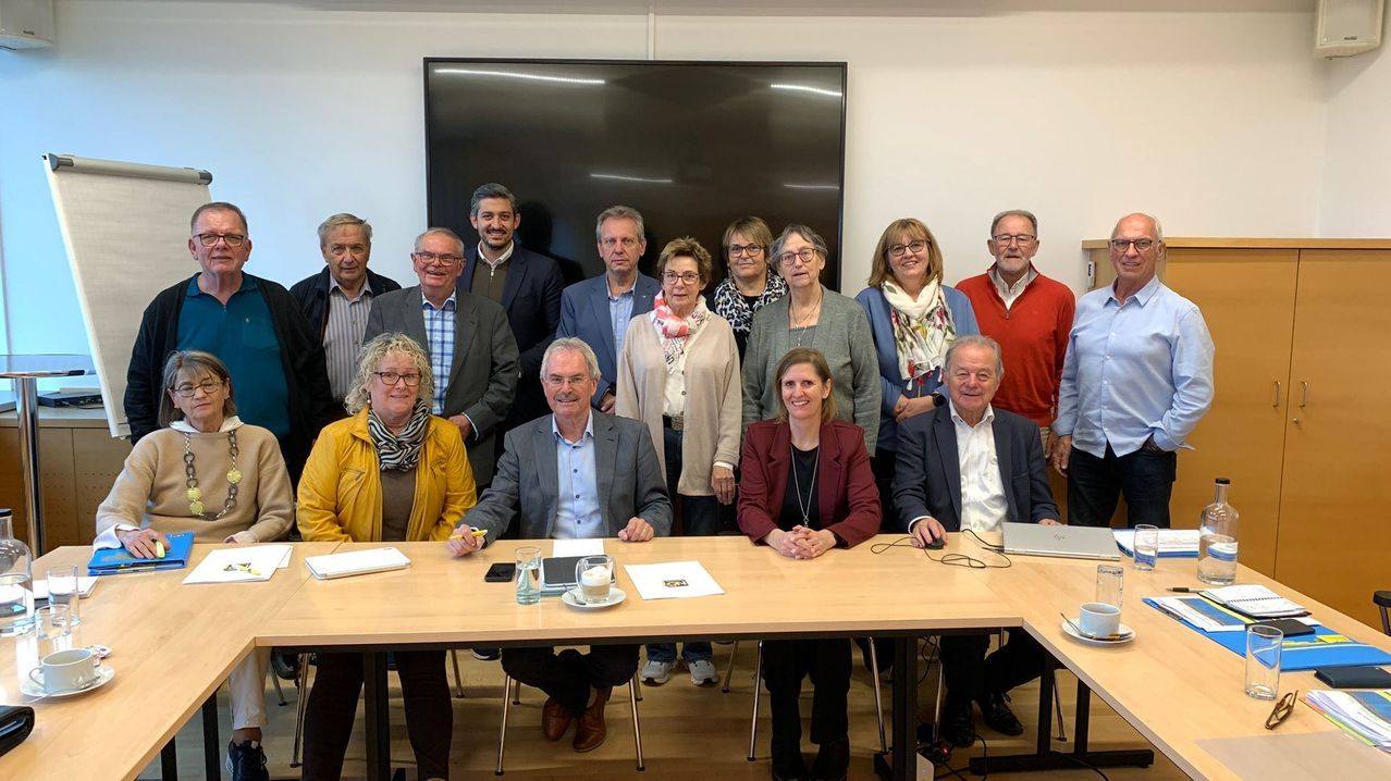 Eine Gruppe von Menschen in formeller Kleidung versammelt sich um einen Tisch, lächelt für ein Foto mit Laptops und Papieren auf dem Tisch. Ein großer Bildschirm ist hinter ihnen.