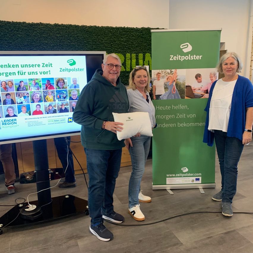 Drei Personen stehen vor einem grünen Banner mit Text. Ein Mann hält ein Kissen. Eine Frau hält ein Papier. Dahinter zeigt ein Monitor eine Collage von Menschen.