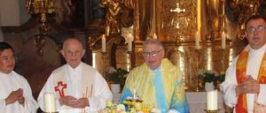 Drei Priester stehen an einem Altar, einer hält ein Buch, ein anderer eine Blumenarrangement, und der dritte hält die Hände gefaltet. Der Altar ist mit Kerzen und Brot geschmückt.