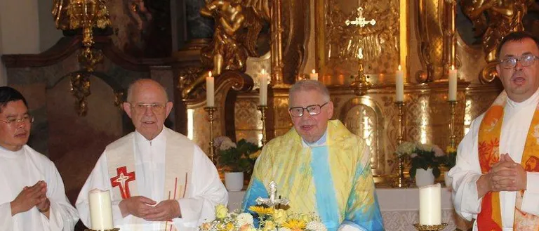 Drei Priester stehen an einem Altar, einer hält ein Buch, ein anderer eine Blumenarrangement, und der dritte hält die Hände gefaltet. Der Altar ist mit Kerzen und Brot geschmückt.