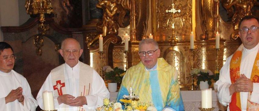 Drei Priester stehen an einem Altar, einer hält ein Buch, ein anderer eine Blumenarrangement, und der dritte hält die Hände gefaltet. Der Altar ist mit Kerzen und Brot geschmückt.
