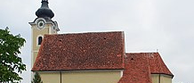 Ein historisches Gebäude mit Glockenturm, rotem Dach und einer Treppe zum Eingang, umgeben von Bäumen und Pflanzen.