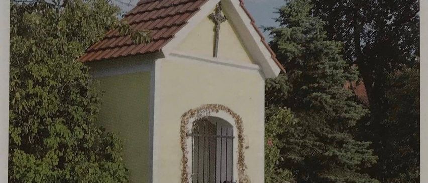 Eine kleine Kapelle mit braunem Dach und einem Kreuz oben, namens Frohlich-Kapelle, befindet sich in Wuschan. Sie hat ein schwarzes Tor und eine Bank in der Nähe.