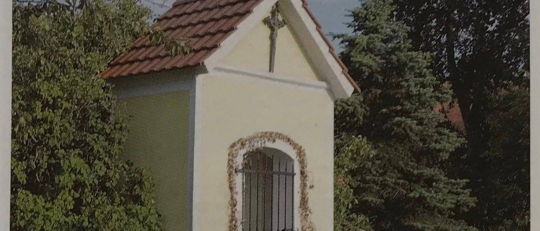 Eine kleine Kapelle mit braunem Dach und einem Kreuz oben, namens Frohlich-Kapelle, befindet sich in Wuschan. Sie hat ein schwarzes Tor und eine Bank in der Nähe.