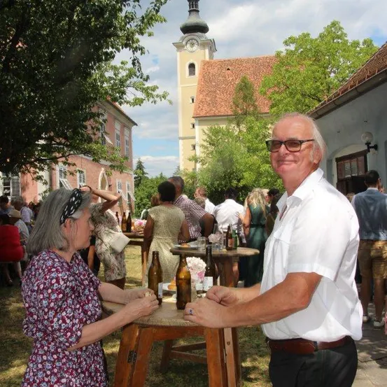 Ein älteres Paar steht lächelnd an einem Tisch mit Flaschen. Dahinter versammelt sich eine Menschenmenge um Tische, und ein Kirchturm ist sichtbar.