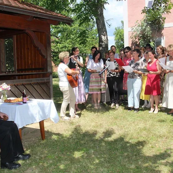 Eine Gruppe von Frauen steht im Kreis und singt, während eine Frau Flöte und eine andere Cello spielt. Ein Tisch mit Brot und einer Flasche ist in der Nähe.