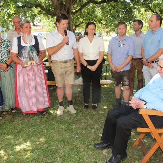 Eine Gruppe von Menschen steht und sitzt unter einem Baum, ein Mann hält ein Mikrofon und spricht, während andere zuhören, eine Frau hält ein Geschenk.