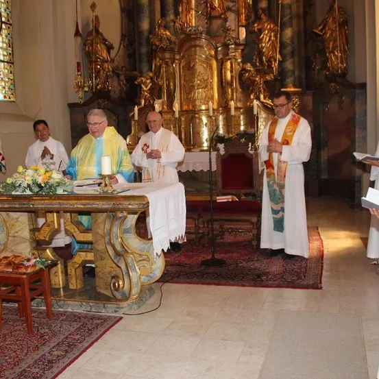 Vier Priester stehen vor einem Altar, möglicherweise während einer religiösen Zeremonie. Der Altar ist mit Blumen und Kerzen geschmückt. Dahinter befindet sich ein goldener Altar mit Statuen und einem roten Teppich.